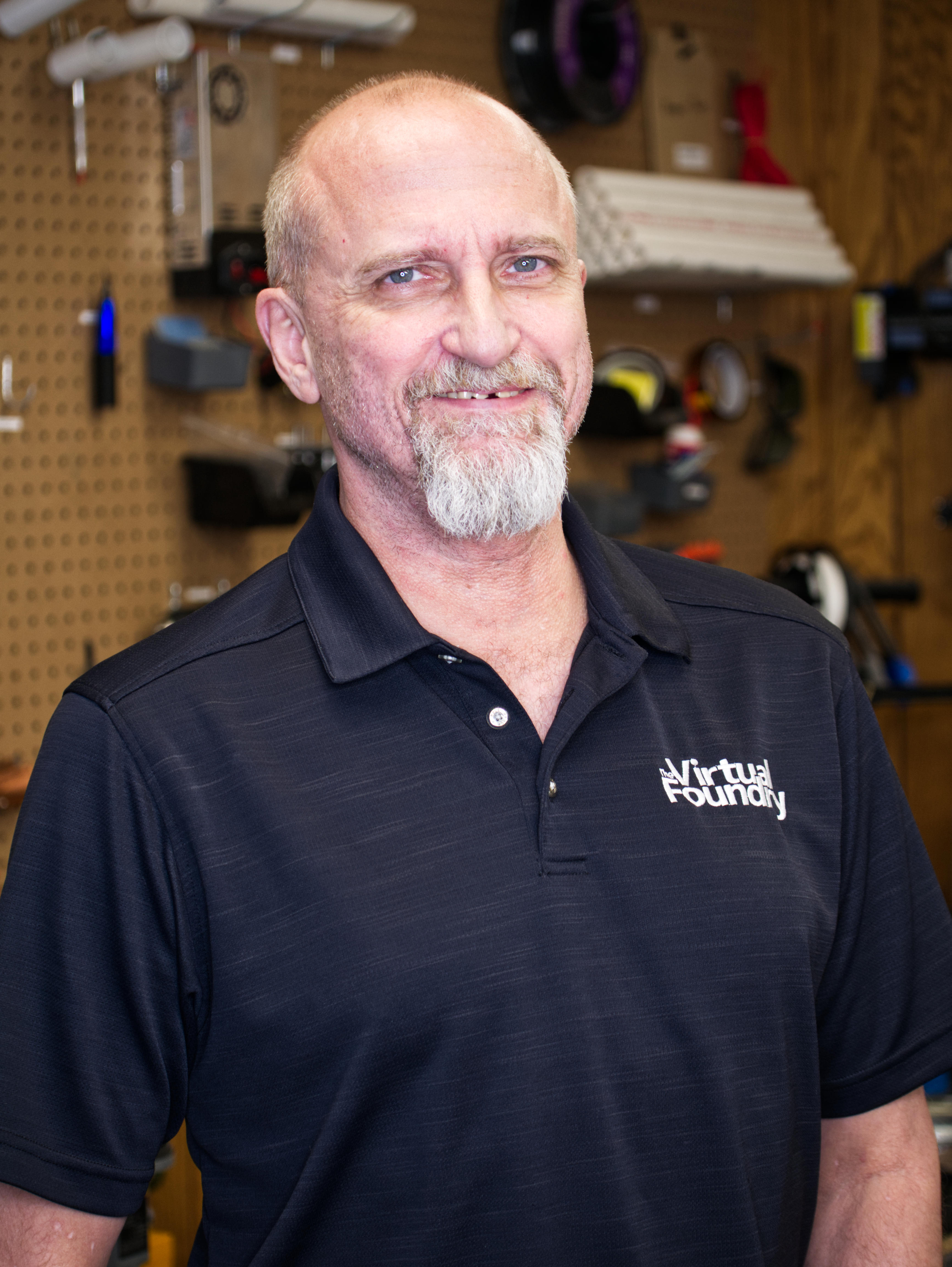 Metal Filament A middle-aged man with a beard smiling at the camera, wearing a black polo shirt with the logo "virtual foundry," standing in a manufacturing workshop.